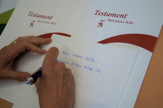 Zwei Hände und Unterlagen für ein Testament. Eine Hand hält einen Stift und schreibt.
