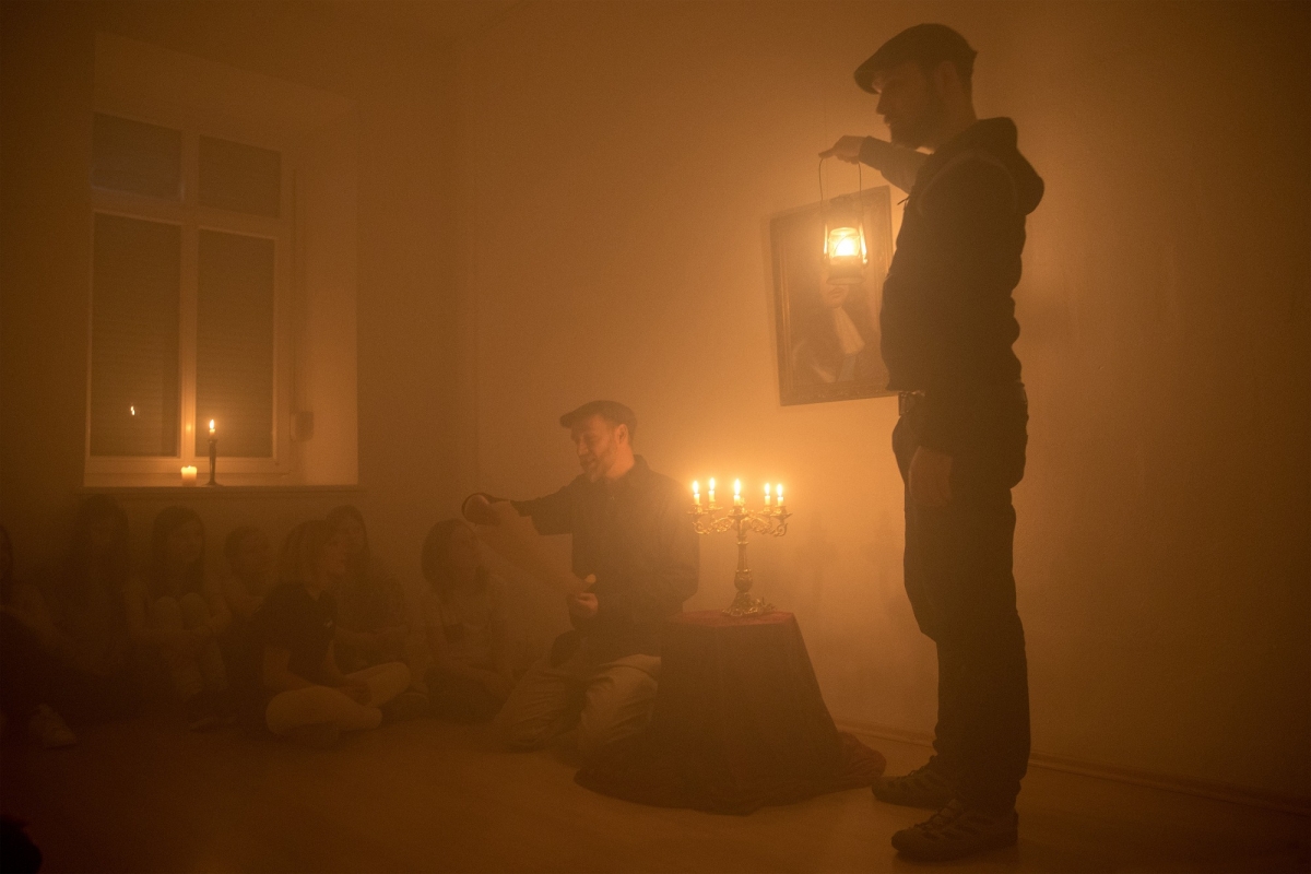 Ein Mann mit Hut steht in einem Zimmer mit Kerzen und Nebel. Er erzählt, auf dem Boden sitzenden Kindern, eine Geschichte.
